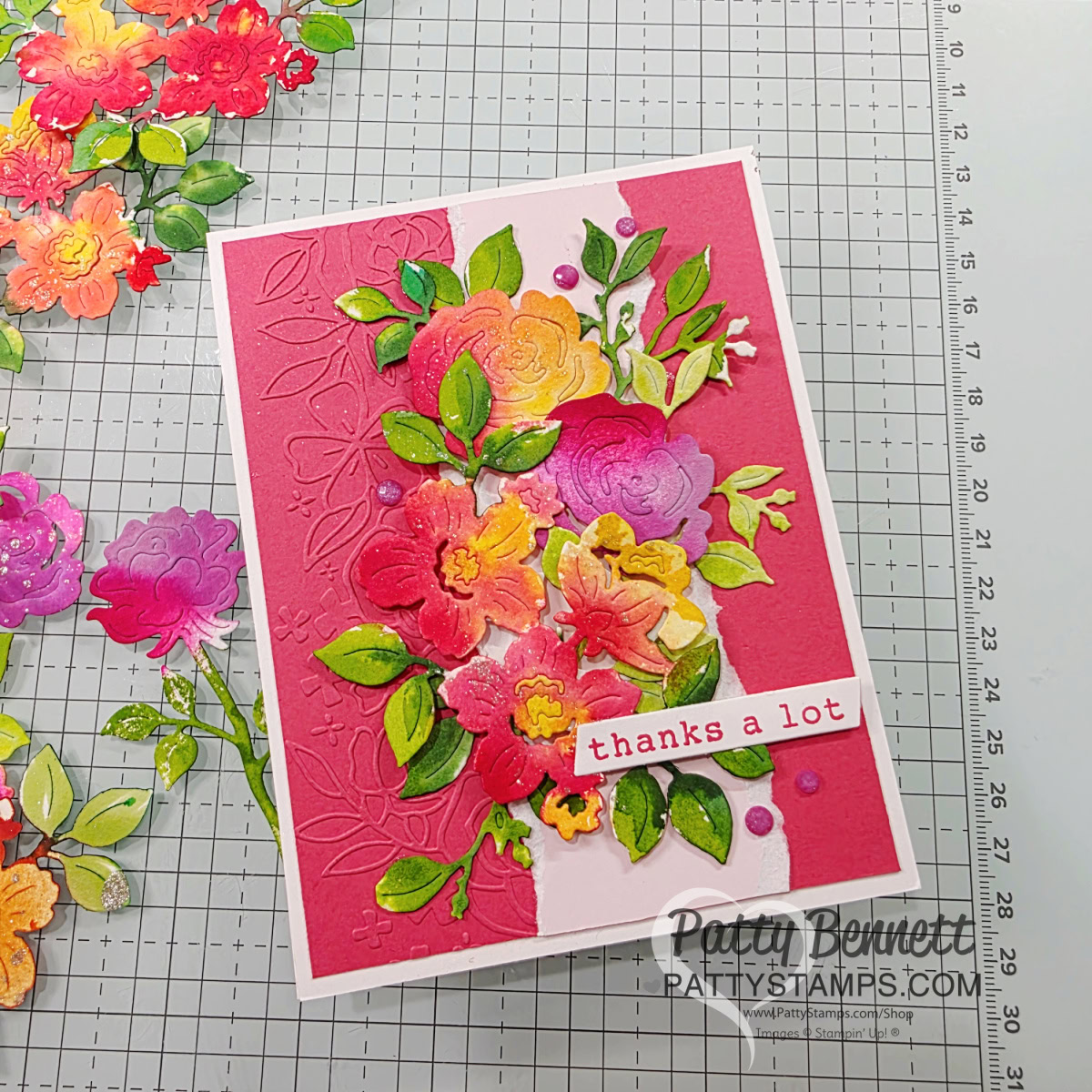 Glass & Gardens embossing folder. A handmade card with layered, colorful paper flowers and green leaves on a textured pink background. The card features a white strip with red text that reads “thanks a lot.” Craft materials and floral cutouts are visible on a gridded work surface. To read more, visit Patty Stamps (pattystamps.com)