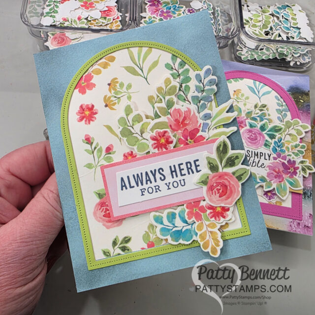 A hand holds a handmade greeting card decorated with colorful watercolor flowers and leaves. The card has an arched frame, green border, and reads “ALWAYS HERE FOR YOU” in bold letters. Other similarly styled cards are visible in the background. To read more, visit Patty Stamps (pattystamps.com)