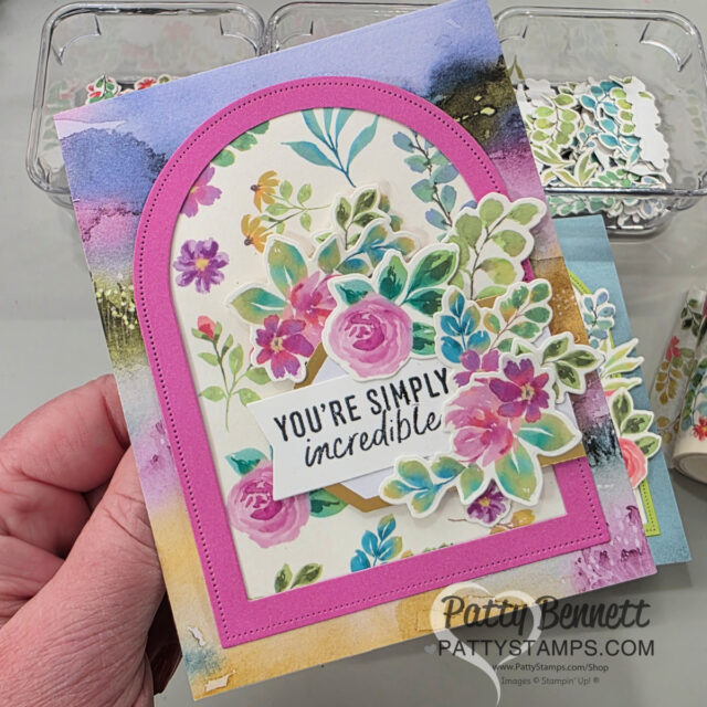 A hand holds a colorful, handmade greeting card decorated with vibrant watercolor-style flowers, green leaves, and a layered pink arched frame. The card features a sentiment: “YOU’RE SIMPLY incredible,” and has a watermark labeled “PattyStamps.com.”. To read more, visit Patty Stamps (pattystamps.com)