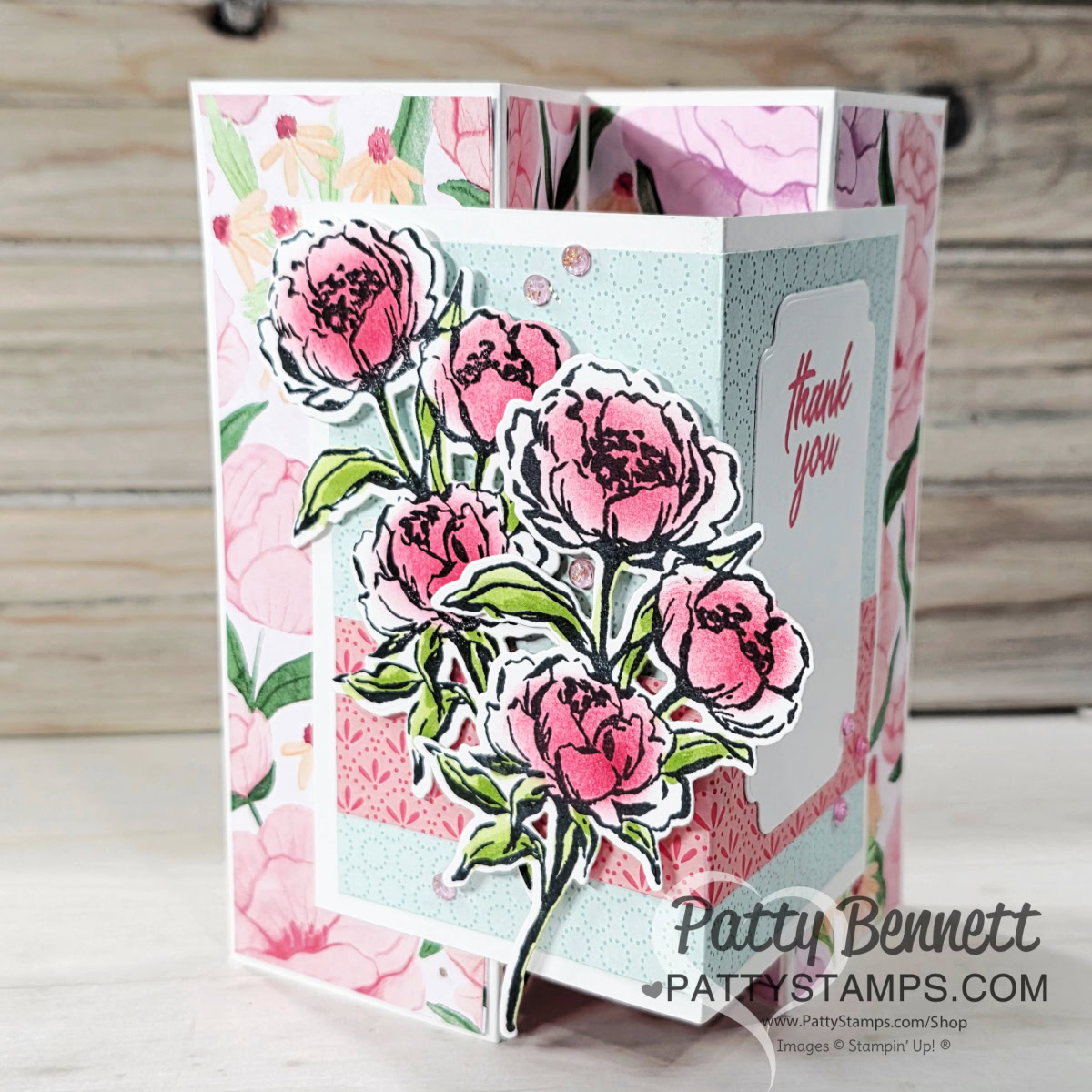 A handmade, three-dimensional thank you card features layered pink peony flowers with green leaves. The card has pastel floral patterns, soft blue and white panels, and a “Thank you” message. Rhinestone embellishments add sparkle. “Patty Bennett” watermark is visible. To read more, visit Patty Stamps (pattystamps.com)