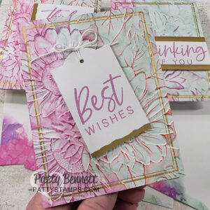 A hand holds a floral-themed greeting card with “Best Wishes” in pink text on a white tag tied to the card. The card has embossed flowers with pastel green and pink hues, gold accents, and is surrounded by similar greeting cards in the background. To read more, visit Patty Stamps (pattystamps.com)