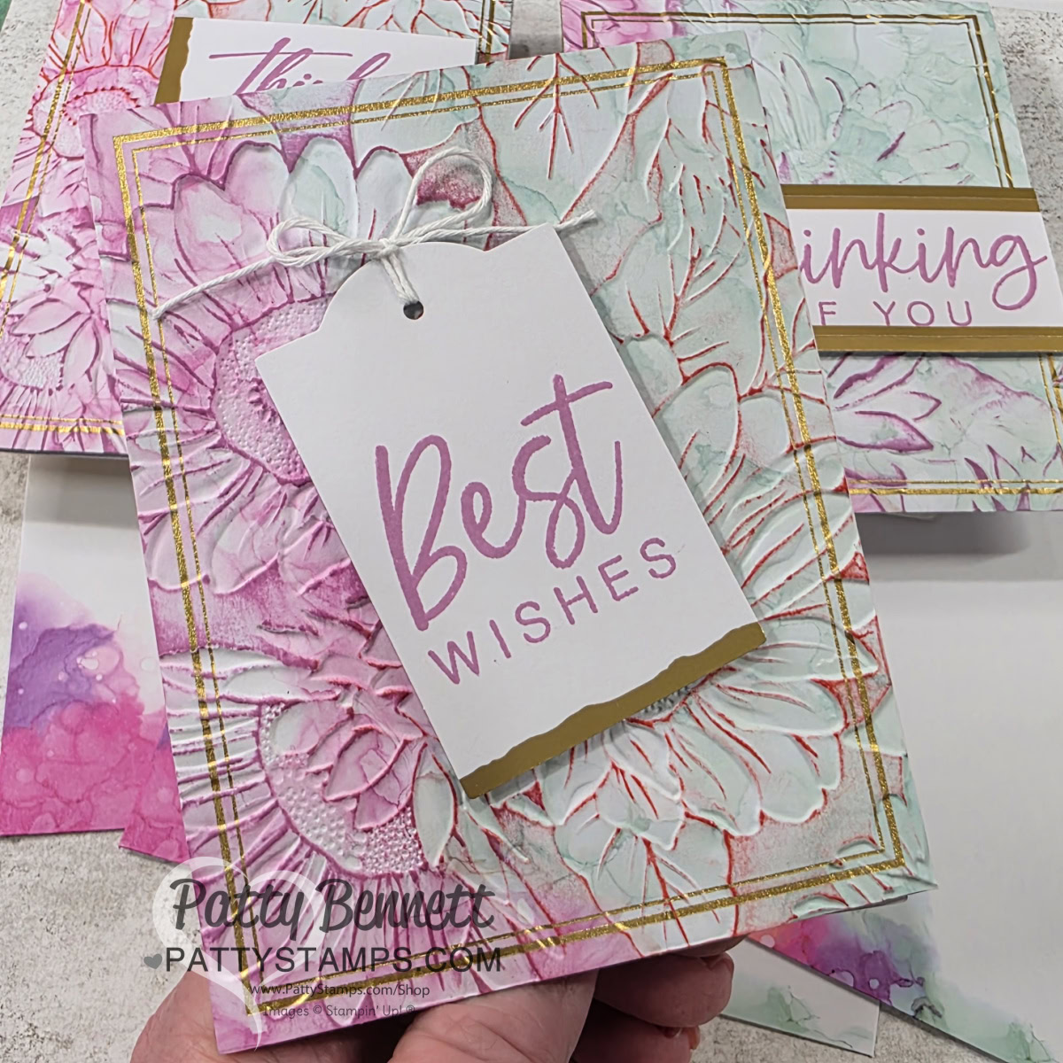 A hand holds a floral-themed greeting card with “Best Wishes” in pink text on a white tag tied to the card. The card has embossed flowers with pastel green and pink hues, gold accents, and is surrounded by similar greeting cards in the background. To read more, visit Patty Stamps (pattystamps.com)