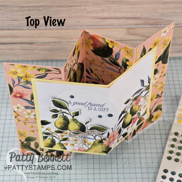 A handmade floral greeting card with a zigzag fold stands open on a grid surface. The card features yellow and pink flowers and green leaves. The front panel displays pears, rhinestone embellishments, and the message "A good friend is a gift. To read more, visit Patty Stamps (pattystamps.com)