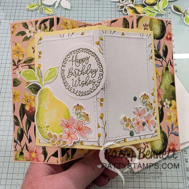A hand holds an open, handmade card from pattystamps, adorned with painterly pears, pink flowers, leaves, and gems. The card features a round "Happy Birthday Wishes" sentiment and a floral background. The watermark reads “Patty Bennett PattyStamps.com.”. To read more, visit Patty Stamps (pattystamps.com)