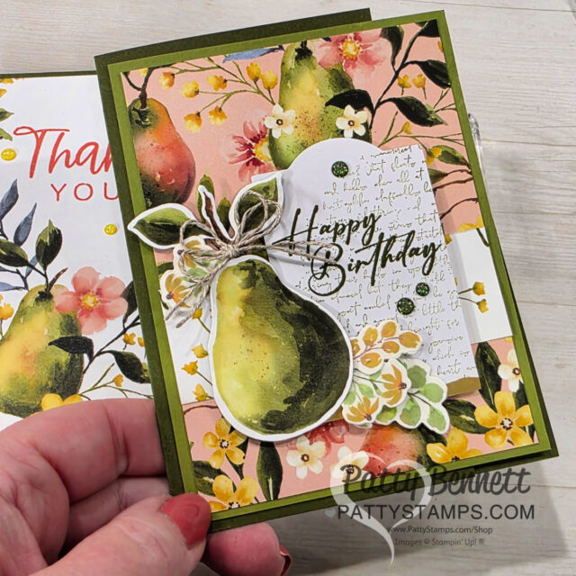A hand holds a floral-themed greeting card decorated with a large watercolor pear, yellow and white flowers, green leaves, and twine. The card has "Happy Birthday" in elegant script on a white cutout, with colorful pear and flower patterns in the background. To read more, visit Patty Stamps (pattystamps.com)