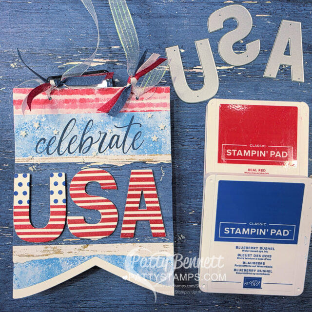 Celebrating the USA banner. A handmade banner reads “celebrate USA” in blue and red letters on a rustic, painted background. Red, white, and blue ribbons are tied at the top. Metal die-cut letters and red and blue Stampin’ Pad ink pads are laid beside the banner on a wooden surface. To read more, visit Patty Stamps (pattystamps.com)
