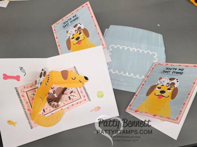 A handmade pop-up card featuring a yellow dog lying down and three kittens sits open on a table, with matching cards and a blue-striped envelope nearby. The cards say, “You’re my best friend” and have dog and cat illustrations on a pink background. To read more, visit Patty Stamps (pattystamps.com)