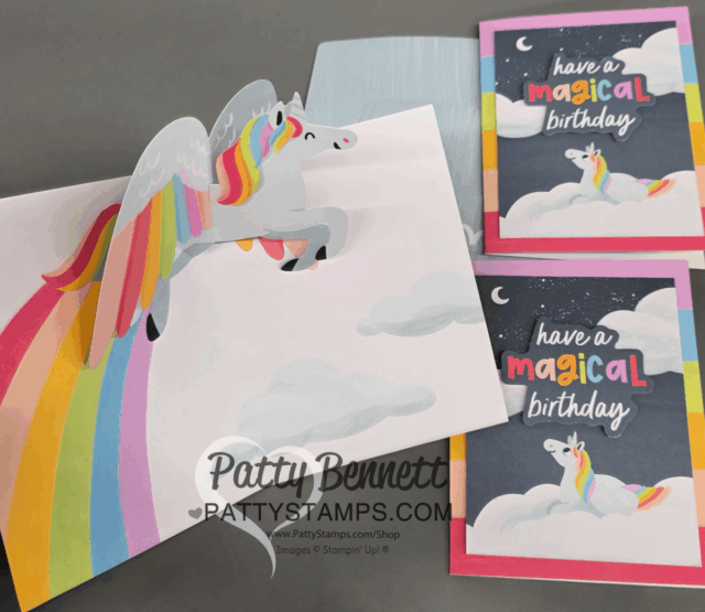 A handmade pop-up card featuring a flying unicorn with a rainbow mane and tail soaring over clouds, with a rainbow trailing behind. Two completed cards next to it display a unicorn, clouds, and the phrase "have a magical birthday" on colorful backgrounds. To read more, visit Patty Stamps (pattystamps.com)