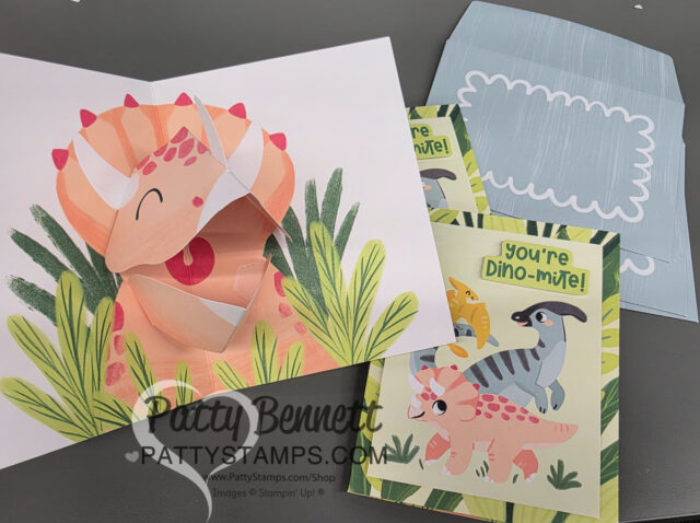 A handmade card set with a dinosaur theme is shown. One card is open, revealing a pop-up pink triceratops among green leaves. Two closed cards display colorful dinosaurs and the text "You're Dino-mite!" An envelope with a blue pattern is in the background. To read more, visit Patty Stamps (pattystamps.com)
