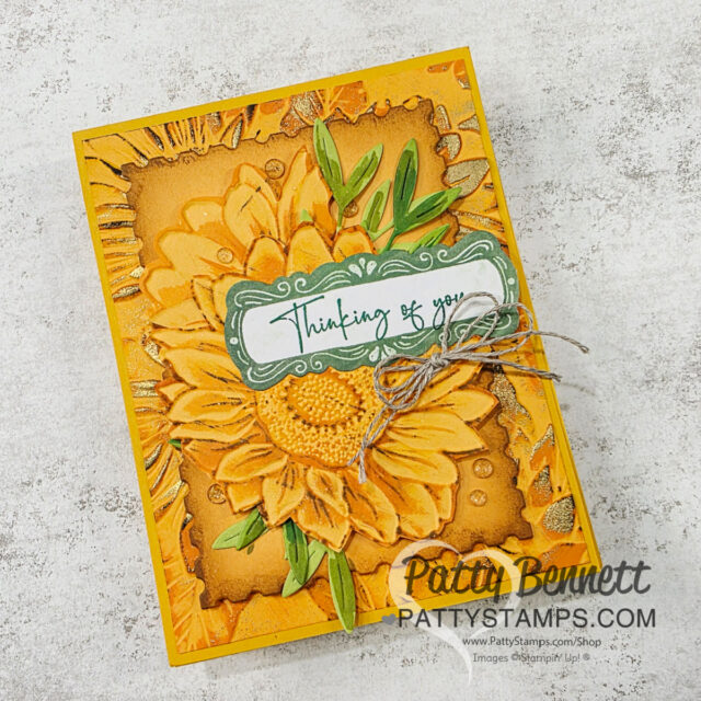 A Very Fancy Sunflower card. Handmade greeting card with a large yellow sunflower on a textured brown and yellow background. Green leaves accent the flower. A green and white label at the center says "Thinking of you." Twine is tied in a bow. “Patty Bennett PattyStamps.com” is at the bottom. To read more, visit Patty Stamps (pattystamps.com)