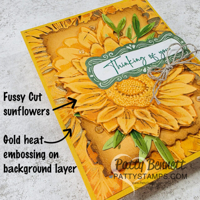 A handmade card features large fussy cut sunflowers with layered petals and green leaves, gold heat embossing on a textured yellow background, a green and white label reading “Thinking of you,” and a twine bow. Text highlights the crafting techniques used. To read more, visit Patty Stamps (pattystamps.com)