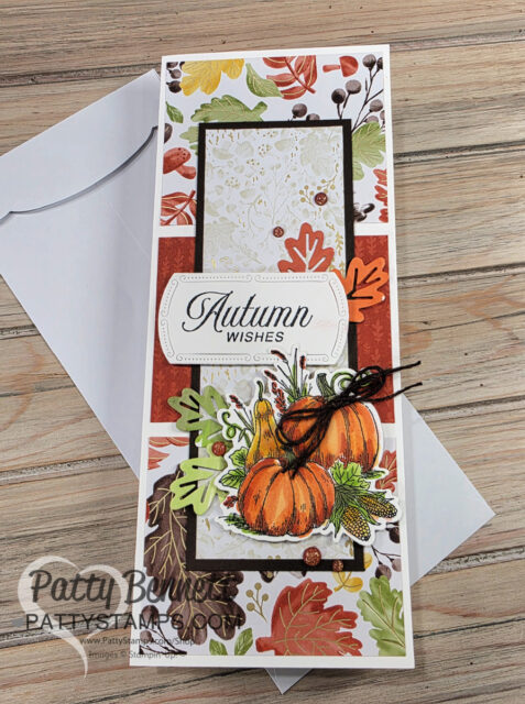 A handmade autumn-themed card features pumpkins, colorful fall leaves, and berries, with “Autumn Wishes” written in elegant script. The card has a collage background in warm tones and is tied with brown twine, resting on a white envelope on a wooden surface. To read more, visit Patty Stamps (pattystamps.com)