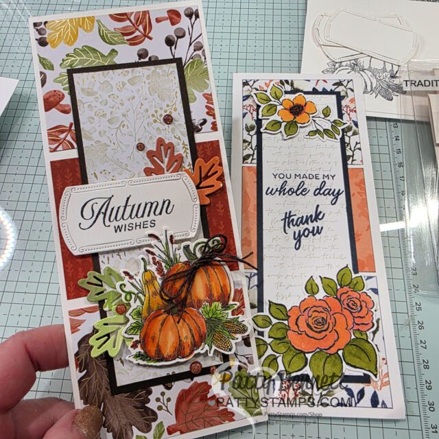 Two tall handmade cards on a craft table: one with pumpkins, leaves, and “Autumn Wishes” text in warm fall colors; the other features orange roses, greenery, and “You made my whole day thank you” text. Clear stamps and crafting supplies are visible in the background. To read more, visit Patty Stamps (pattystamps.com)