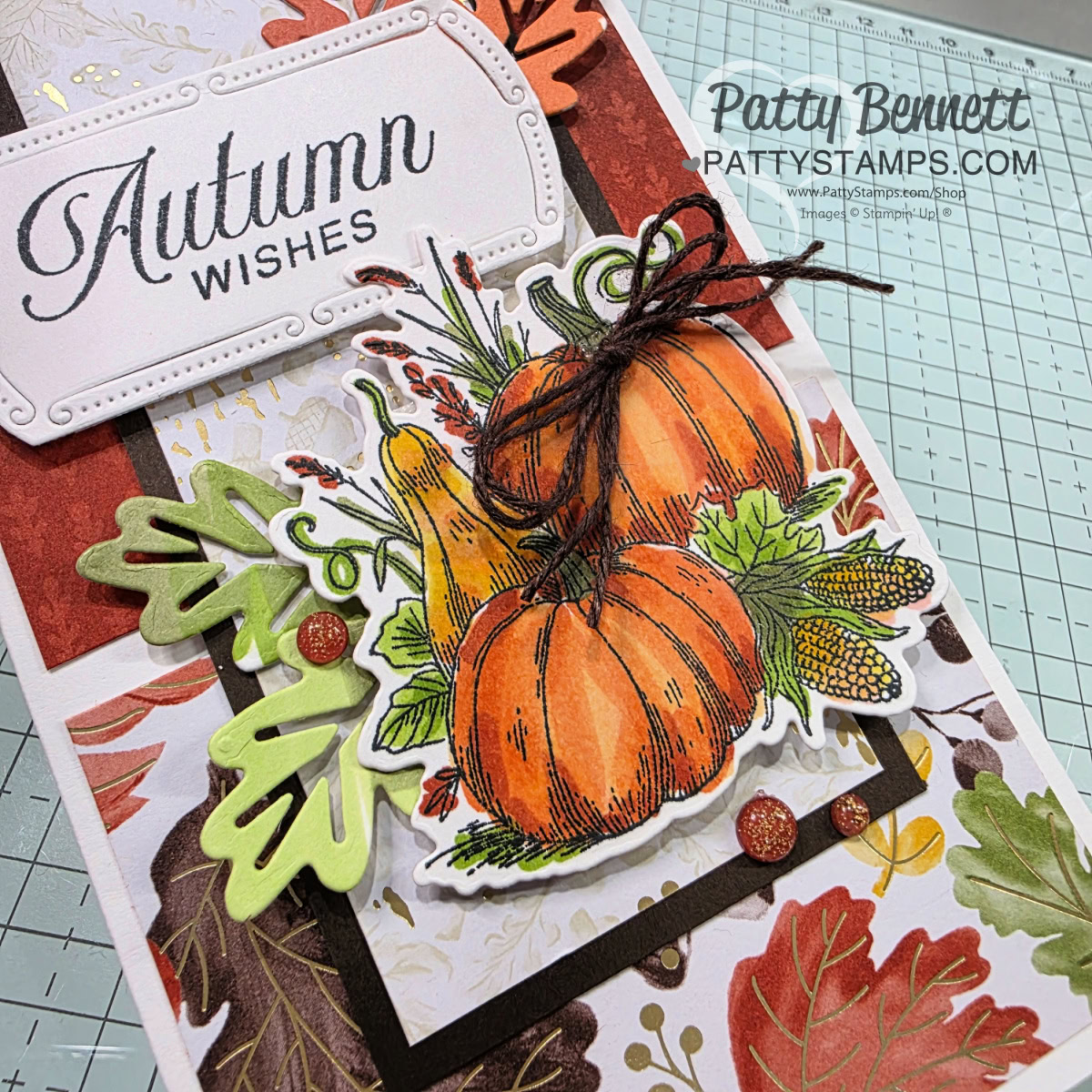 A handmade card decorated with stamped and colored pumpkins, leaves, and a tied brown string. The card says "Autumn Wishes" on a white label. Leaf and dot embellishments and layered, autumn-themed papers add texture and detail to the card. To read more, visit Patty Stamps (pattystamps.com)