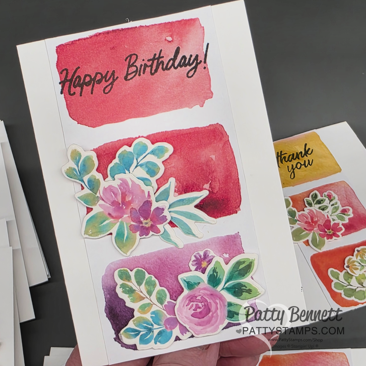 A hand holds a greeting card decorated with three watercolor brushstroke backgrounds in red and pink tones, each topped with floral stickers and the words "Happy Birthday!" in black script at the top. Other colorful cards are partially visible in the background. To read more, visit Patty Stamps (pattystamps.com)