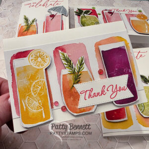 A handmade card features three colorful watercolor drinks with lemon slices and garnishes, and a stitched white rectangle with “Thank You” in red script. The card is held in hand, with more similarly styled cards visible in the background. “Patty Bennett” is printed below. To read more, visit Patty Stamps (pattystamps.com)