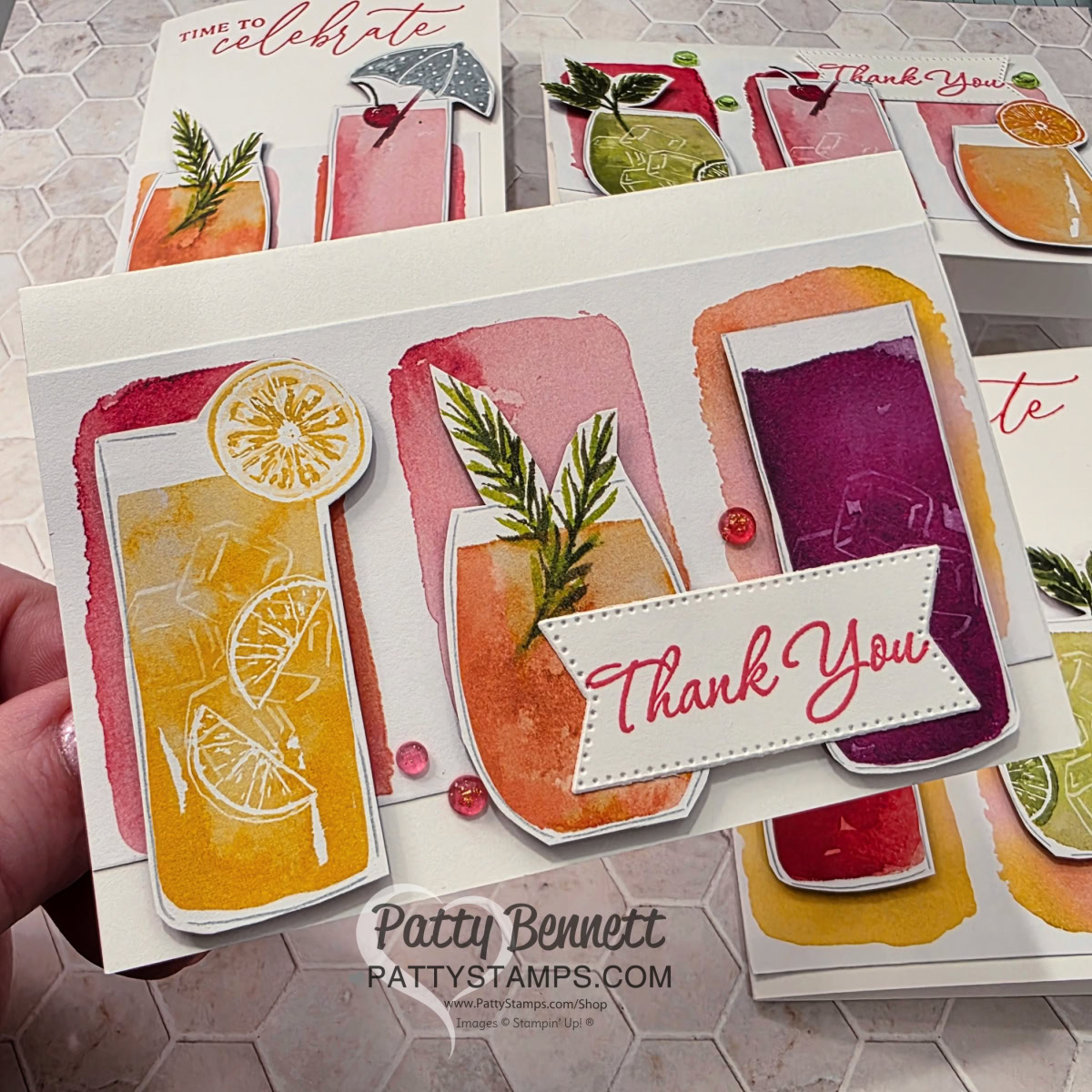 A handmade card features three colorful watercolor drinks with lemon slices and garnishes, and a stitched white rectangle with “Thank You” in red script. The card is held in hand, with more similarly styled cards visible in the background. “Patty Bennett” is printed below. To read more, visit Patty Stamps (pattystamps.com)