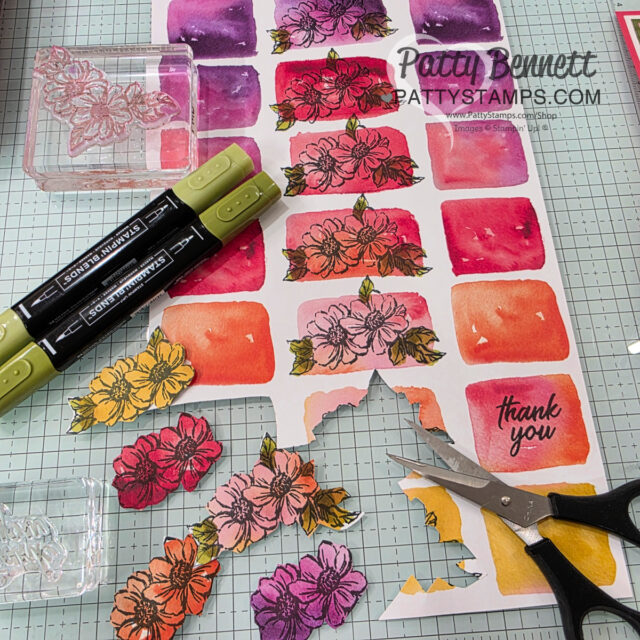 A crafting scene with colorful watercolor swatches, floral stamp images, markers, small scissors, and a “thank you” card. Cut-out stamped flowers in pink, red, yellow, and purple are arranged on a gridded surface. The watermark reads "Patty Bennett PATTYSTAMPS.COM. To read more, visit Patty Stamps (pattystamps.com)