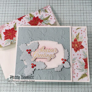A handmade holiday card features embossed light blue paper with holly leaves and red berries, a “Seasons Greetings” label in gold, and poinsettia-themed border and envelope. Card rests on a grid background with “Patty Bennett pattystamps.com” watermark. To read more, visit Patty Stamps (pattystamps.com)
