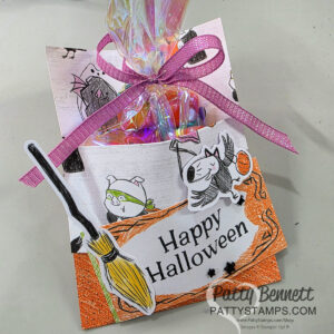 A handmade Halloween treat holder decorated with playful cat and bat drawings, a broom cutout, and an orange textured border. It holds colorful candy in a clear bag tied with a purple ribbon. "Happy Halloween" is written on the front. To read more, visit Patty Stamps (pattystamps.com)