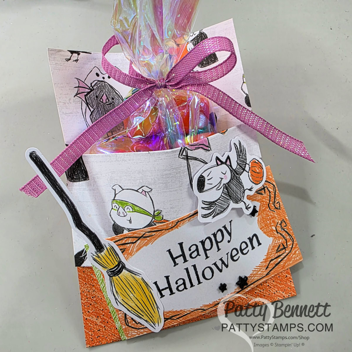 A handmade Halloween treat holder decorated with playful cat and bat drawings, a broom cutout, and an orange textured border. It holds colorful candy in a clear bag tied with a purple ribbon. "Happy Halloween" is written on the front. To read more, visit Patty Stamps (pattystamps.com)