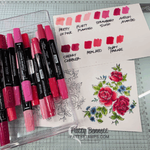 A clear box holds pink and red Stampin’ Blends markers beside a floral card partially colored with vibrant pinks and greens. A swatch card shows different marker shades labeled "Pretty in Pink," "Flirty Flamingo," "Strawberry Slush," "Melon Mambo," "Cherry Cobbler," "Real Red," and "Poppy Parade. To read more, visit Patty Stamps (pattystamps.com)