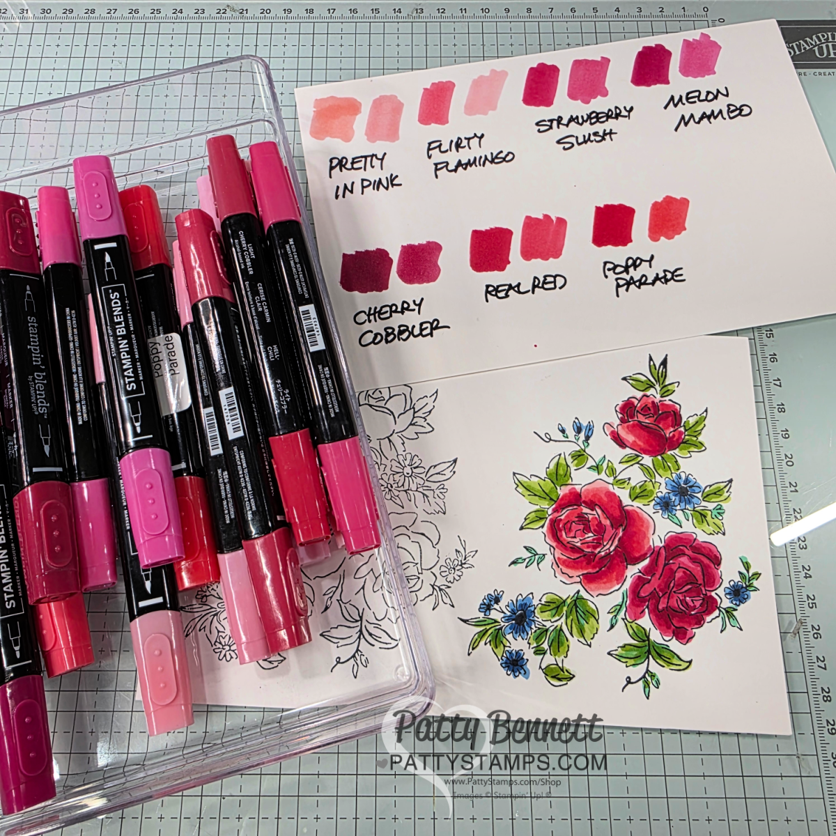 A clear box holds pink and red Stampin’ Blends markers beside a floral card partially colored with vibrant pinks and greens. A swatch card shows different marker shades labeled "Pretty in Pink," "Flirty Flamingo," "Strawberry Slush," "Melon Mambo," "Cherry Cobbler," "Real Red," and "Poppy Parade. To read more, visit Patty Stamps (pattystamps.com)