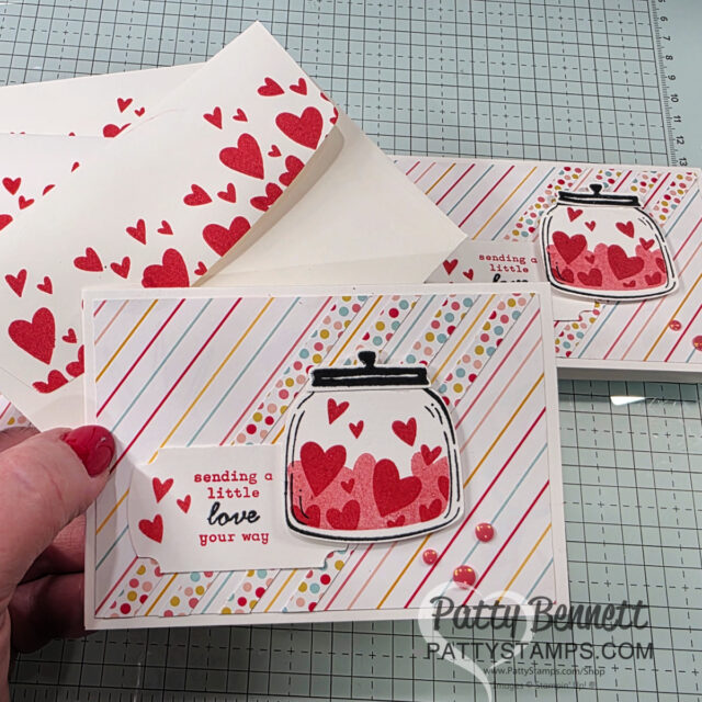 A handmade card features a jar filled with red hearts on a pastel-striped background. "Sending a little love your way" is stamped on the front. Matching envelopes with red hearts are in the background. The card is held by a hand, with two more cards below. To read more, visit Patty Stamps (pattystamps.com)