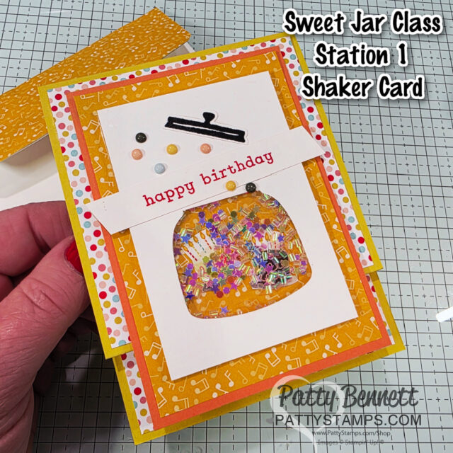 A handmade birthday shaker card features a clear jar filled with colorful sequins, a “happy birthday” banner, and a bright dotted border. The card rests on a grid mat and is held by a hand; text reads “Sweet Jar Class Station 1 Shaker Card.”. To read more, visit Patty Stamps (pattystamps.com)