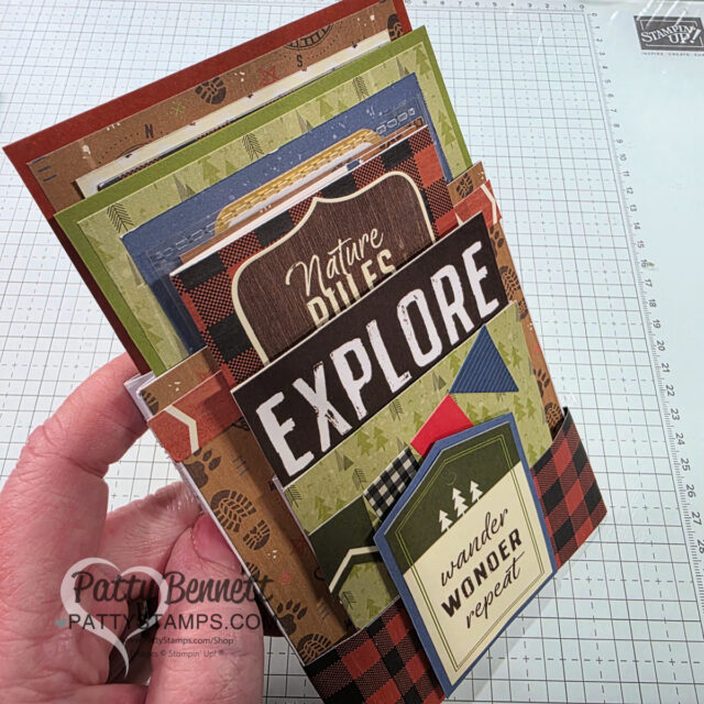 A hand holds a layered, nature-themed scrapbook with cards and tags. The front displays the words "EXPLORE" and "wander WONDER repeat." Earthy colors and patterns feature trees, plaid, and leaves. A crafting mat and ruler are visible in the background. To read more, visit Patty Stamps (pattystamps.com)