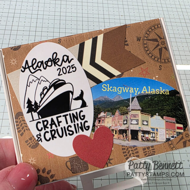A handmade card features layered brown and tan paper, a red heart, a “Skagway, Alaska” label with a town photo, and a white oval with a cruise ship, mountains, and text: “Alaska 2025 Crafting & Cruising.” A hand holds the card over a grid surface. To read more, visit Patty Stamps (pattystamps.com)