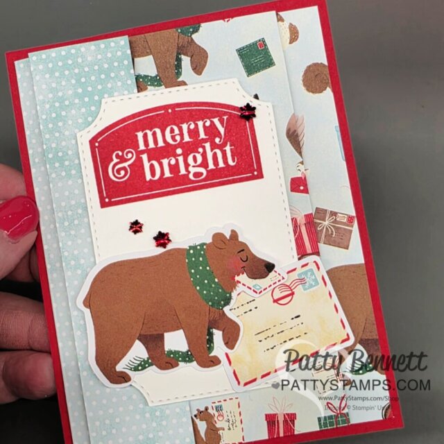 A handmade holiday card features a large brown bear carrying a letter, set against festive bear and gift-themed paper. A bold red label reads “merry & bright.” Three small red star embellishments add sparkle. “Patty Bennett - PattyStamps.com” is printed at the bottom. To read more, visit Patty Stamps (pattystamps.com)