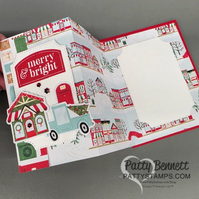Hand holding an open, colorful Christmas card decorated with festive houses, a green truck carrying a tree, and a candy shop. The card features the text “merry & bright” and has a blank white space for a message. Patty Bennett’s watermark is at the bottom. To read more, visit Patty Stamps (pattystamps.com)