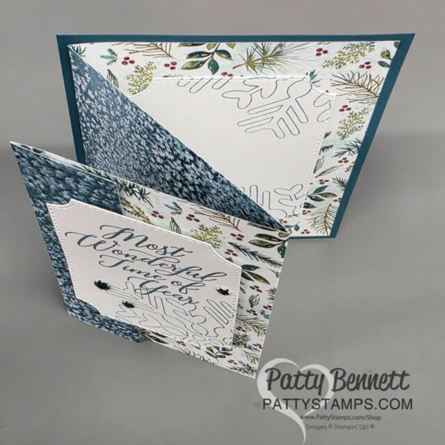 A handmade winter-themed greeting card is displayed standing open, showing layered patterned papers with pine branches, snowflakes, and blue textured designs. A square front panel reads “Most Wonderful Time of the Year” in cursive. The card has a zig-zag fold. To read more, visit Patty Stamps (pattystamps.com)