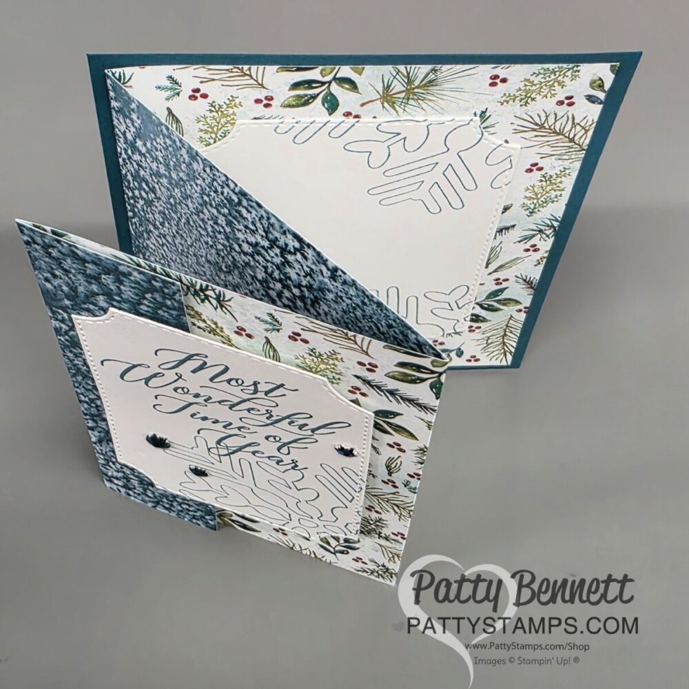 A handmade winter-themed greeting card is displayed standing open, showing layered patterned papers with pine branches, snowflakes, and blue textured designs. A square front panel reads “Most Wonderful Time of the Year” in cursive. The card has a zig-zag fold. To read more, visit Patty Stamps (pattystamps.com)