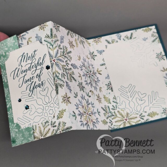 A handmade holiday card is open, revealing winter-themed patterned paper with snowflakes and pine branches. The left panel features a tag reading “Most Wonderful Time of the Year” in script. White die-cut snowflakes decorate both panels. "PattyStamps.com" is printed on the card. To read more, visit Patty Stamps (pattystamps.com)