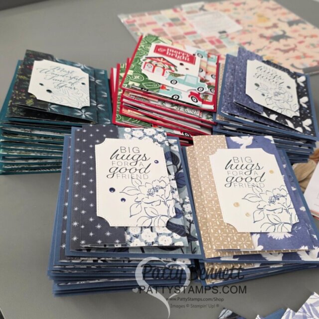 Stacks of handmade greeting cards with decorative fronts are arranged on a table. Each card features a tag reading “BIG hugs FOR A good FRIEND,” surrounded by different patterned paper designs in blue, gold, red, and green. To read more, visit Patty Stamps (pattystamps.com)