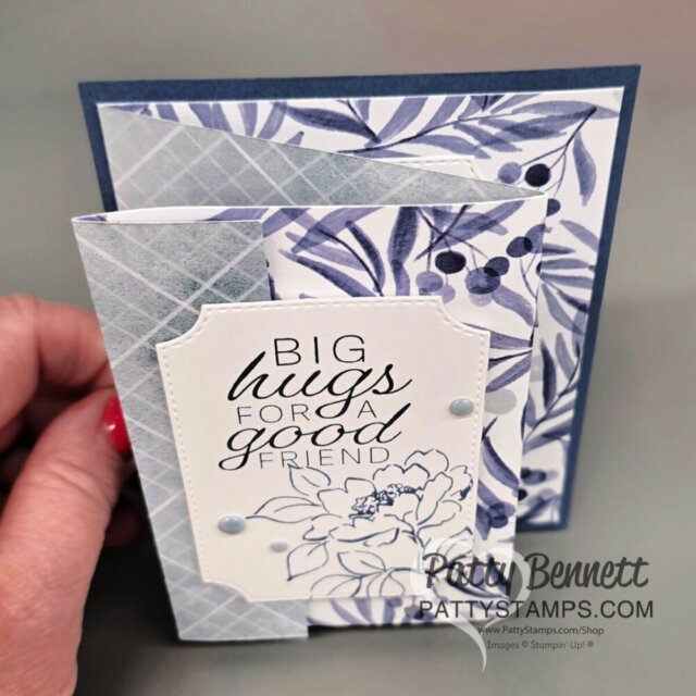A hand holds a handmade greeting card with a blue and white floral theme. The card has layered panels with leafy and geometric patterns, and a front label that reads, "BIG hugs FOR A good FRIEND," adorned with a floral outline and decorative dots. To read more, visit Patty Stamps (pattystamps.com)