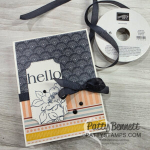 Handmade greeting card with “hello” in black script on a white label featuring a floral outline. The card has navy floral patterned paper on top and orange-yellow stripes below, accented with black ribbon, a bow, and black beads. A Stampin’ Up! ribbon spool sits nearby. To read more, visit Patty Stamps (pattystamps.com)
