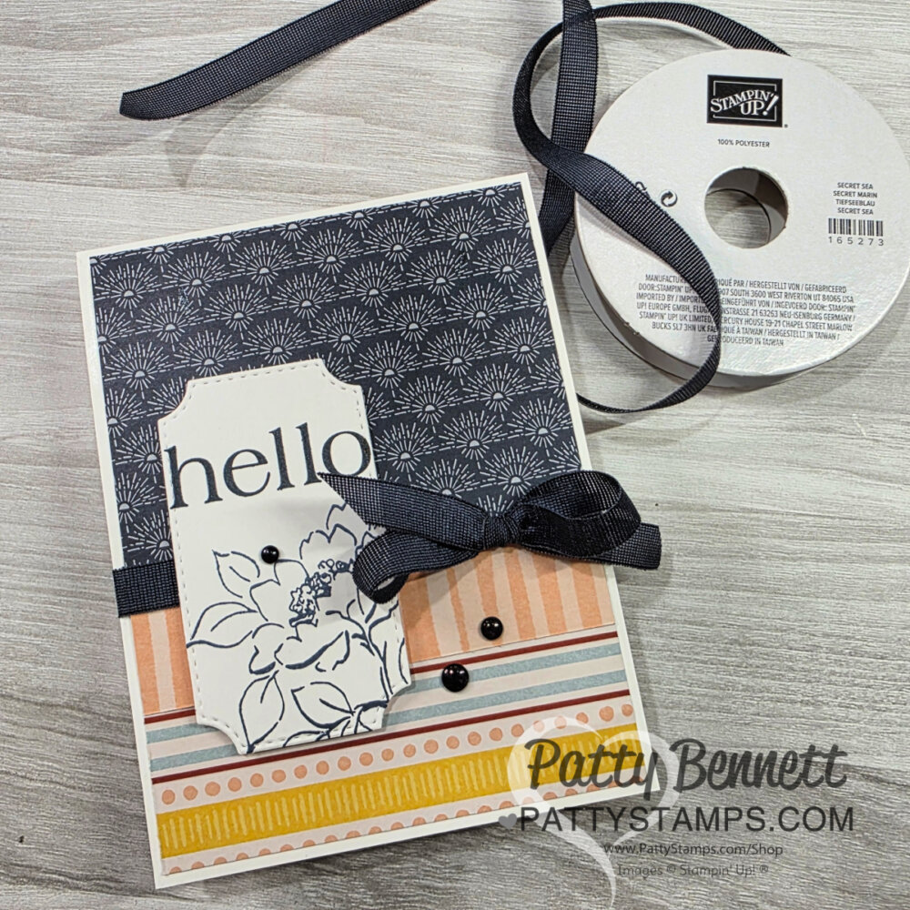 Handmade greeting card with “hello” in black script on a white label featuring a floral outline. The card has navy floral patterned paper on top and orange-yellow stripes below, accented with black ribbon, a bow, and black beads. A Stampin’ Up! ribbon spool sits nearby. To read more, visit Patty Stamps (pattystamps.com)