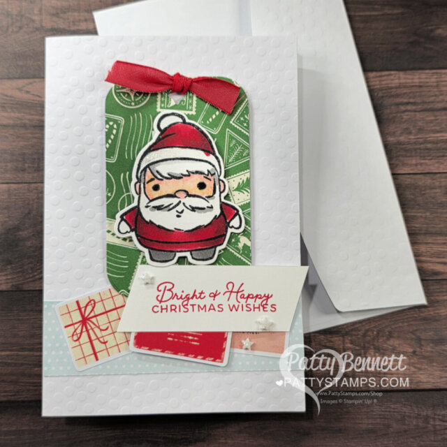 A handmade Christmas card features a cute, cartoon Santa on a green patterned tag with a red ribbon. The card is white with embossed dots and includes a “Bright & Happy Christmas Wishes” message and small gift-wrapped images in the bottom corner. To read more, visit Patty Stamps (pattystamps.com)