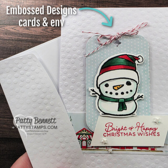 A hand holding a white card and envelope with raised dot embossing. The card features a cute snowman tag with a red-and-white string, and a “Bright & Happy Christmas Wishes” message. “Embossed Designs cards & env” is written at the top. To read more, visit Patty Stamps (pattystamps.com)