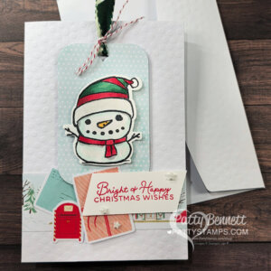A handmade Christmas card with a textured white background features a tag with a smiling snowman in a red and green hat and scarf. Below are pastel postcards, a red mailbox, stars, and a message: "Bright & Happy CHRISTMAS WISHES." An envelope sits behind. To read more, visit Patty Stamps (pattystamps.com)