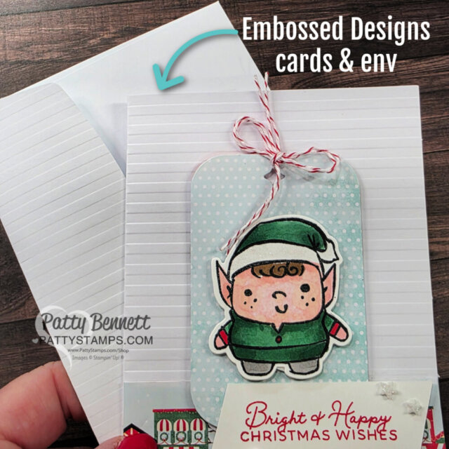 A hand holds a white embossed greeting card with matching envelope. On the card is a cute tag featuring a cartoon elf in green, tied with red-and-white string. Text on the image reads “Embossed Designs cards & env” with a blue arrow pointing to the card and envelope. To read more, visit Patty Stamps (pattystamps.com)
