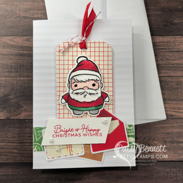 A handmade Christmas card with a cute cartoon Santa on a red grid tag, topped with a red ribbon. Below Santa are layered cards and a white label reading "Bright & Happy CHRISTMAS WISHES" in red. The card background is white and textured. To read more, visit Patty Stamps (pattystamps.com)