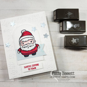 A handmade Christmas card features a cute, cartoon Santa on a white embossed background with festive words. "Santas Coming to Town" is stamped in red below Santa. Three star embellishments accent the card. Nearby are three brown paper punches and a "Patty Bennett" watermark. To read more, visit Patty Stamps (pattystamps.com)
