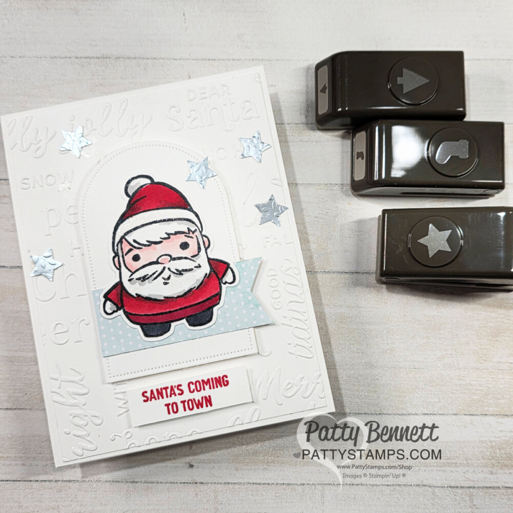 A handmade Christmas card features a cute, cartoon Santa on a white embossed background with festive words. "Santas Coming to Town" is stamped in red below Santa. Three star embellishments accent the card. Nearby are three brown paper punches and a "Patty Bennett" watermark. To read more, visit Patty Stamps (pattystamps.com)