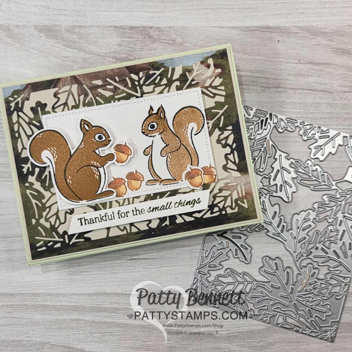 A handmade card features two cartoon squirrels holding acorns with the phrase “Thankful for the small things.” The card has a leafy border layer and sits next to a silver metal leaf-shaped die on a wood grain surface. "PattyStamps.com" is watermarked below. To read more, visit Patty Stamps (pattystamps.com)