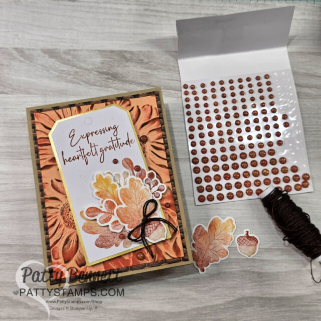 A handmade card with autumn leaves and “Expressing heartfelt gratitude” text is on a wood surface. Next to it are brown embellishment dots, brown string, an acorn sticker, and leaf cut-outs. The card has warm orange, yellow, and brown tones. To read more, visit Patty Stamps (pattystamps.com)