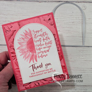 A hand holds a pink, handmade card with an embossed floral background. The card features a large pink sunflower, uplifting script text, and a thank you message for support during a battle. A metal die-cut arch shape lies on the wooden surface beside the card. To read more, visit Patty Stamps (pattystamps.com)